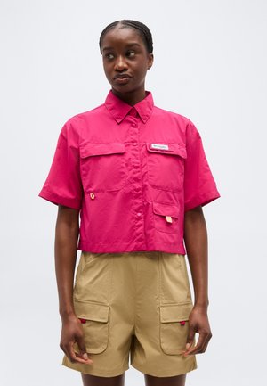 Γυναίκα που φοράει φωτεινό ροζ πουκάμισο με κοντά μανίκια και κουμπιά, με μπροστινές τσέπες, και μπεζ cargo σορτς, μπροστά σε απλό φόντο.