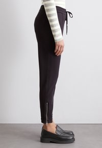 Zwarte joggerbroeken met elastische taille en koord, voorzien van zijzakken en ritsen bij de enkels, gemaakt van een gladde stof.
