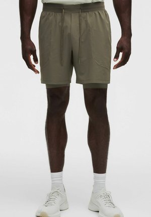 Mann trägt olivgrüne, geschichtete Shorts mit Kordelzug, weiße Socken und weiße Sportschuhe, steht vor einem einfarbigen Hintergrund.