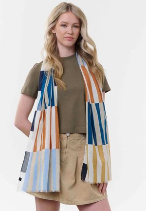 Junge Frau mit langen blonden Haaren trägt ein olivgrünes T-Shirt, einen beigen Rock und einen mehrfarbigen, gestreiften Schal, der um ihren Hals drapiert ist.