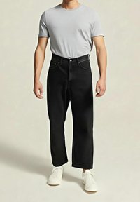 Adulte portant une chemise à manches courtes gris clair, un pantalon noir ample et des chaussures blanches sans lacets, debout devant un fond beige uni.