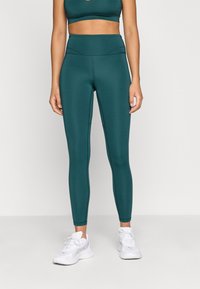 Türkiz színű, magas derekú leggings sima textúrával, egy hozzá illő crop toppal párosítva. Finom varrásokkal díszítve, fehér sportcipővel viselve.