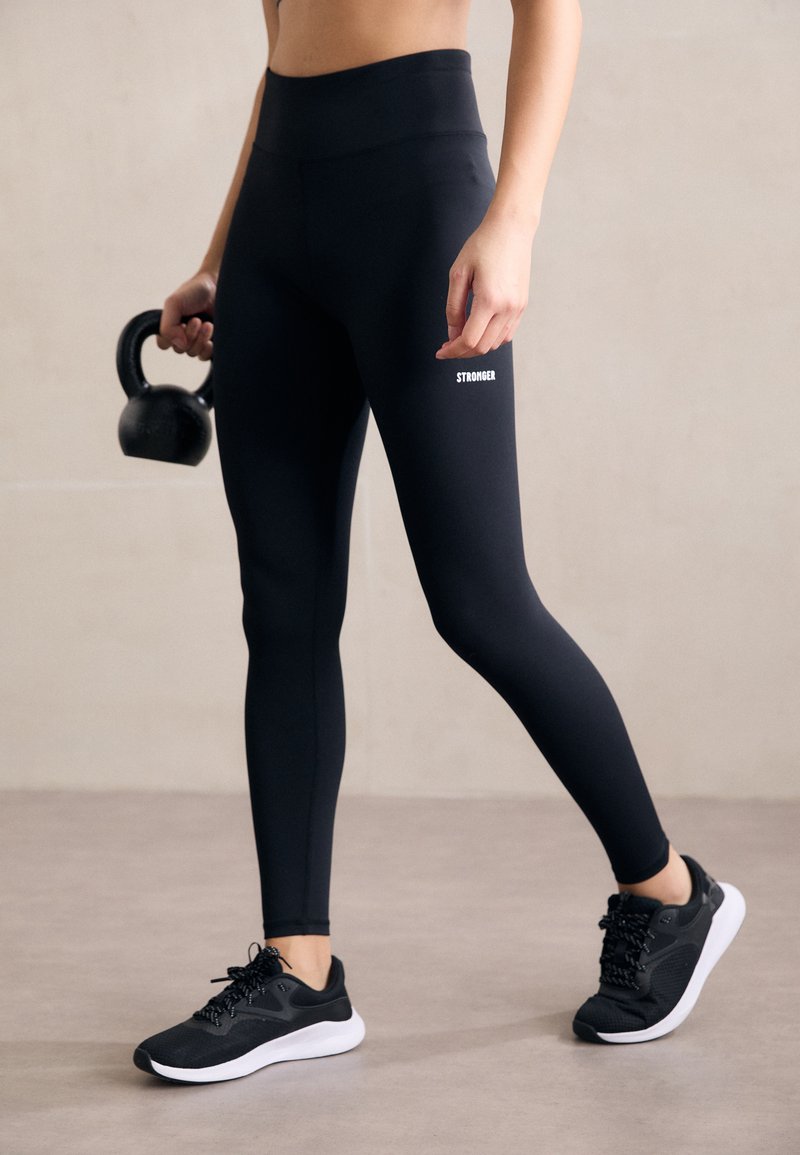 Black high-waisted leggings with a smooth texture and a white "STRONGER" logo on the side, paired with black athletic shoes and a kettlebell.