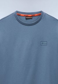 Camiseta de algodón azul con cuello redondo acanalado y un logo en relieve con forma de bandera; cuenta con una etiqueta de logo naranja en el interior del cuello.