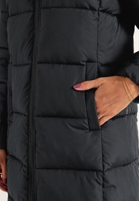 Svart pufferjacka med en strukturerad, quiltad design, dragkedja fram och sidofickor. Slät, isolerad tyg med en glänsande yta.