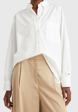 Person wearing loose white button-up shirt with front pocket and beige high-waist wide-leg trousers against plain background.