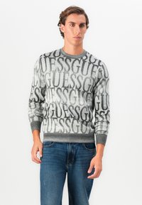 Maglione grigio con grande logo "GUESS" ripetuto in tonalità a contrasto. Textura a maglia con polsini e orlo a coste. Indossato con jeans blu.