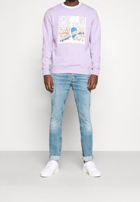 Lila sweatshirt med grafiskt texttryck i vitt och orange, ihopparad med ljusblå jeans och vita sneakers, stående mot en neutral bakgrund.