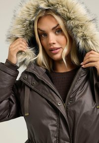 Veste isolante marron avec une capuche en fausse fourrure. Présente une fermeture éclair et à bouton-pression, des poignets élastiques et une doublure intérieure texturée.