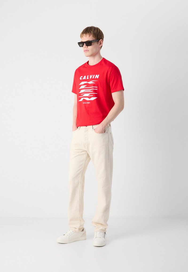 T-shirt en coton rouge avec texte et graphisme blancs, pantalon crème et baskets blanches. Modèle portant des lunettes de soleil noires, debout devant un fond uni.