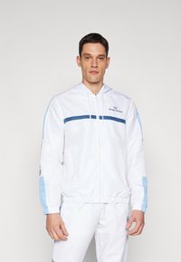 Chaqueta blanca con cremallera y acentos en azul claro, con capucha. Presenta una franja horizontal azul en el pecho y puños elásticos.