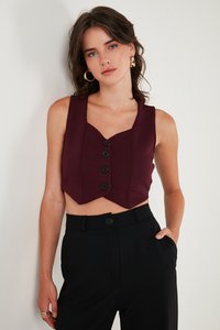 Mujer con top corto sin mangas color burdeos con botones y pantalones negros de talle alto, de pie con una mano en el bolsillo contra una pared blanca.