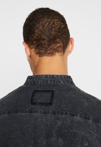 Camicia in denim nera con colletto, caratterizzata da un dettaglio ricamato a forma di quadrato sulla schiena. Il tessuto ha un aspetto leggermente testurizzato.