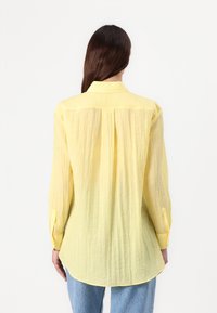 Camicia button-up gialla, con texture, dotata di yoke posteriore e maniche lunghe, con i polsini arrotolati e un orlo curvo.