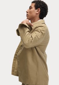 Beige katoenen jas met een kraaghalslijn, een voorzak en knoopsluitingen. De stof heeft een gladde textuur en een relaxte pasvorm.