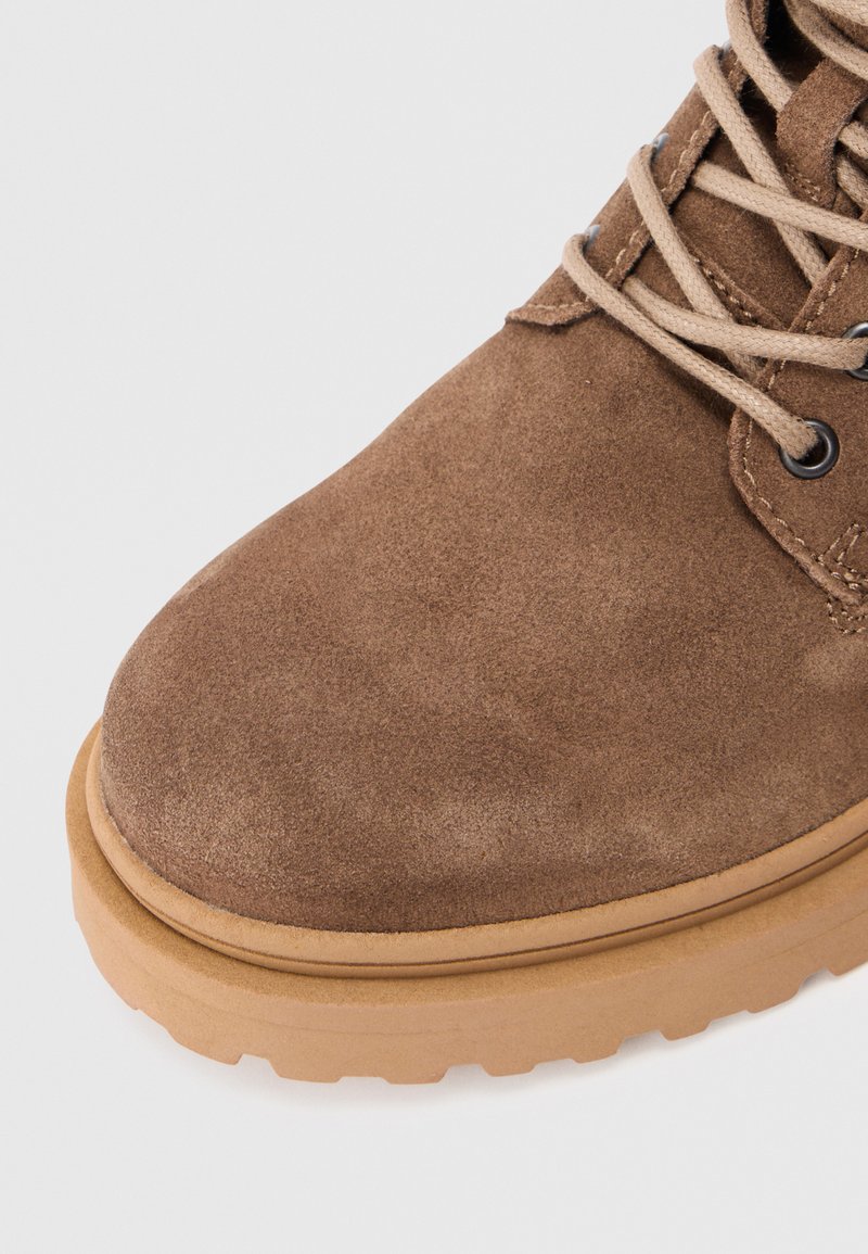 Brown suede boot with a round toe, contrasting tan rubber sole, and central lace-up design with fabric laces and metal eyelets.