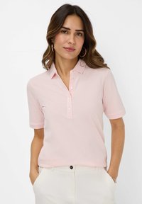 Lys pink polo t-shirt med korte ærmer, en knaplukning foran og struktureret stof. Bæres med hvide bukser, hænderne i lommerne.