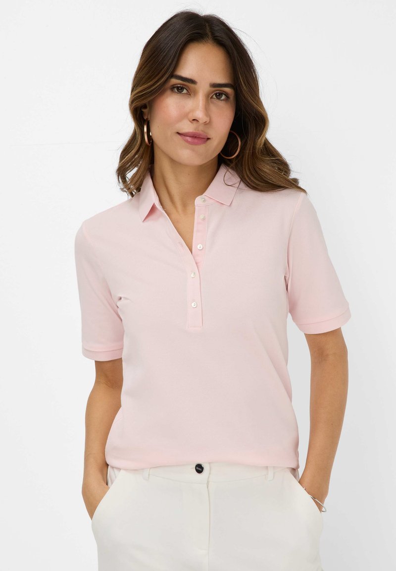 Lys pink polo t-shirt med korte ærmer, en knaplukning foran og struktureret stof. Bæres med hvide bukser, hænderne i lommerne.