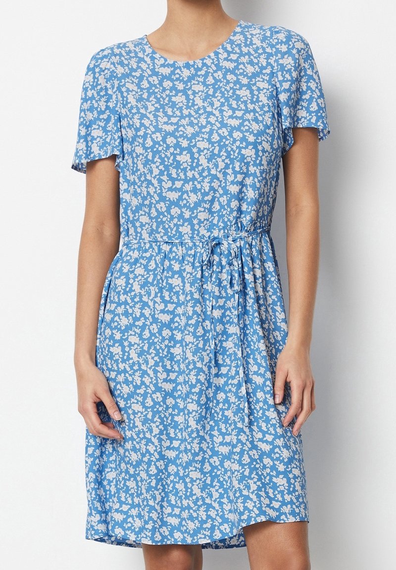 Žena v modrých šatech s krátkým rukávem a bílým květinovým vzorem, se stahovacími šňůrkami v pase, na jednobarevném pozadí.