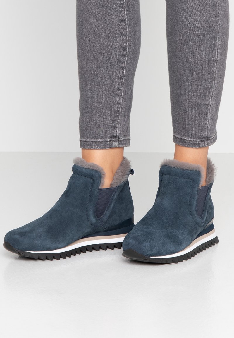 Bottes bleu en daim montant à la cheville avec doublure en fourrure grise, panneaux élastiques sur les côtés, semelles noires rainurées portées avec un jean gris raccourci.