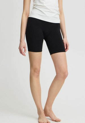 Femme portant un short noir moulant mi-cuisse et un haut blanc sans manches, debout pieds nus devant un fond uni.
