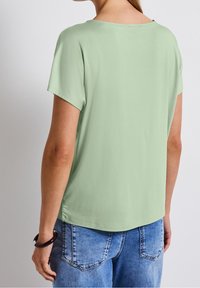 Mintgrön kortärmad topp med avslappnad passform, slät textur, rundad halsringning och en något längre bakre nederkant, tillsammans med ljusblå jeans.