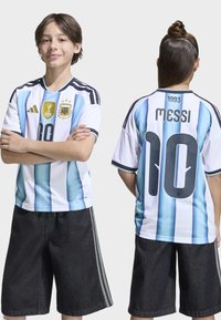 Dwóch chłopców w koszulkach piłkarskich Argentyny, jeden zwrócony przodem z założonymi rękami, a drugi pokazujący plecy z napisem "MESSI 10" oraz rokiem 1893.