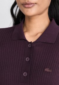 Ribana purpurna polo majica s klasičnim kragnom, četiri dugmeta i malim izvezenim logotipom na lijevoj strani grudi.