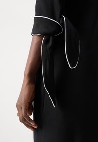 Black dress with short sleeves featuring white piping and tied accents. Smooth fabric texture; hand partially shown for scale.