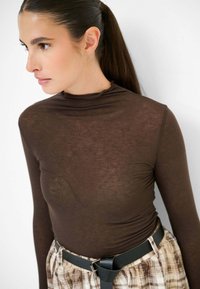 Maglia lunga marrone con maniche lunghe realizzata in tessuto sottile e testurizzato, con colletto alto, abbinata a una gonna a quadri e una cintura nera.