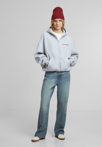 Haut à capuche zippé gris avec le mot "PROHIBÉ" en rouge, associé à un jean en denim bleu clair et un bonnet en tricot bordeaux. Coupe décontractée, deux poches.