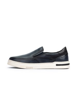Zapato negro de cuero tipo slip-on con un acabado suave, punta redondeada, suela blanca en contraste y detalles de diseño minimalistas.