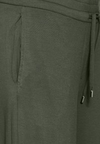 Ciemnozielone spodnie dresowe wykonane z miękkiego materiału, wyposażone w boczne kieszenie oraz elastyczny pas z regulacją na sznurku z metalowymi akcentami. Gładka tekstura.