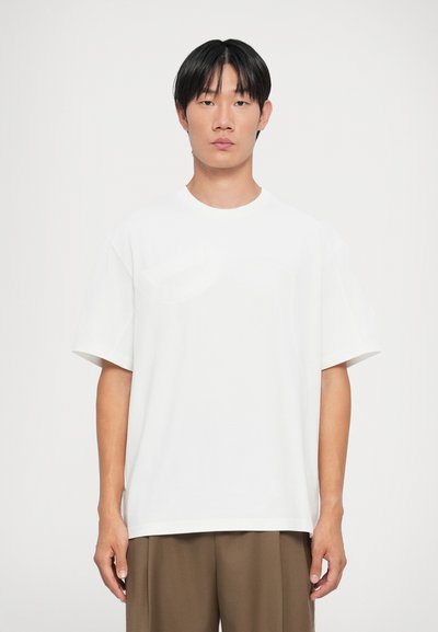 T-shirt blanc à manches courtes avec un col rond et une poche sur la poitrine gauche, fabriqué dans un tissu lisse et léger.
