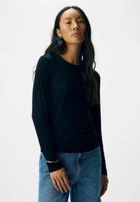 Sort langærmet skjorte med rund halsudskæring, lavet af et glat stof. Sammen med lyseblå højtaljede jeans og et simpelt sølvarmbånd.
