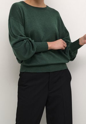 Personne portant un pull vert foncé à manches longues avec des manches bouffantes et un pantalon noir, ajustant la manche d'une main sur un fond uni.
