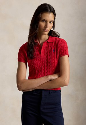 Γυναίκα με μακριά σκούρα μαλλιά που φοράει κόκκινη κοντομάνικη πλεκτή πόλο μπλούζα και σκούρα παντελόνια, στέκεται με τα χέρια σταυρωμένα μπροστά σε ουδέτερο φόντο.