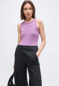 Jeune femme portant un haut lilas sans manches et un pantalon noir taille haute à jambes larges, tenant un sac à main noir froncé.