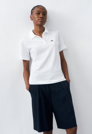 Donna che indossa una polo bianca con un piccolo logo e pantaloncini neri fino al ginocchio, in piedi con le mani in tasca davanti a una parete bianca.