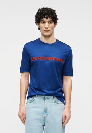 Homme aux cheveux bouclés foncés portant un t-shirt bleu Emporio Armani et un jean bleu clair, debout devant un fond uni.