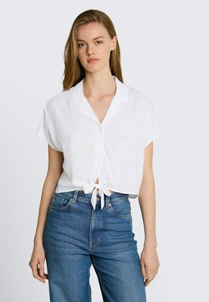 Frau trägt ein weißes kurzärmliges Hemd, das in der Taille gebunden ist, und hoch taillierte blaue Jeans, steht vor einem schlichten Hintergrund.