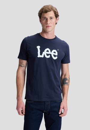 Joven con tatuajes que lleva una camiseta azul marino de Lee y vaqueros oscuros, de pie frente a un fondo liso de color gris claro.