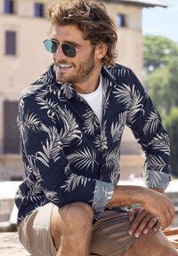 Korte mouwen, marineblauw shirt met een beige palmbladmotief, met een knoopsluiting kraag en lichtblauwe manchetten. Draagt over een wit T-shirt en beige shorts.