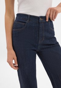 Granatowe dżinsy o wysokim stanie, z kieszeniami z przodu i widocznym przeszyciem. Materiał jest gładki z lekko teksturowanym wykończeniem.