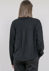 Chemise noire à manches longues avec une finition texturée, présentant une couture de yoke dans le dos et une coupe décontractée. Le tissu semble léger et tombe doucement.