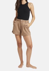 Musta hihaton tanktoppi, yhdistettynä korkeavyötäröisiin, vaaleanbeigeihin, laskostettuihin shortseihin, joissa on sivutaskut ja nappisuljenta.