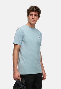 Ljusblå bomulls t-shirt med korta ärmar, rund halsringning och en liten mörk logotyp på vänster bröst. Modellen håller en mönstrad väska.