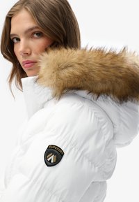 Chaqueta tipo puffer blanca con capucha de piel sintética marrón extraíble, presenta un parche negro con el logo 'Superdry' en la manga y textura acolchada.