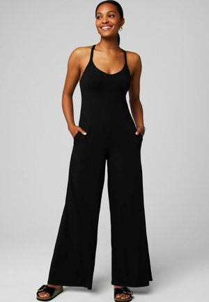 Woman wearing a sleeveless black jumpsuit with wide legs, hands in pockets, smiling and looking to the side against a gray background.