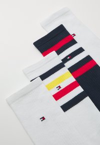 Tre par med sokker: én hvit, én med røde, marineblå og gule striper, og et stripete mønster med hvite, røde, marineblå og gule detaljer.